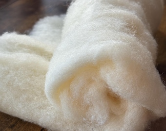 May include: A soft, off-white roll of raw wool fiber with a fluffy, fibrous texture. The material is loosely coiled, showcasing the natural, unspun state of the white wool. The fibers appear dense and airy, forming a continuous, thick layer.