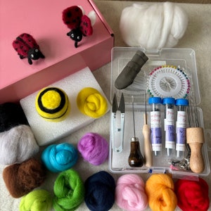 May include: A needle felting craft kit featuring a pink box, two felted ladybugs, and various colored wool roving bundles. A clear plastic organizer case contains metal needles, a wooden handle, scissors, a leather finger guard, and pins.