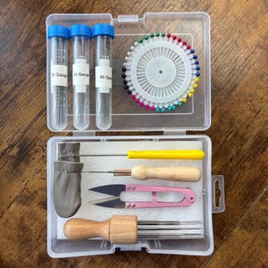 May include: A clear plastic craft supply organizer containing three clear tubes with blue caps labeled 36, 38, and 40 Gauge, a circular pin cushion with multi-colored ball-head pins, a leather finger guard, a yellow tool, a wooden tool, pink snips, and a multi-needle tool.