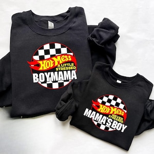 Pode incluir: Duas camisolas pretas com os dizeres "Hot Mess Boymama" e "Hot Mess Mama's Boy" em branco, vermelho e amarelo. O design inclui um círculo quadriculado e chamas.