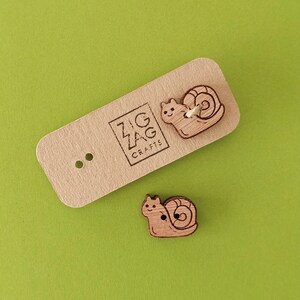 May include: Two wooden snail buttons with a cute cartoon face. The buttons are on a light brown card with the text "Zig Zag Crafts" printed on it.