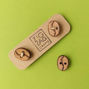 Puede incluir: Tres botones de madera con forma de granos de café con un acabado de madera marrón. Los botones tienen un agujero en el centro y están etiquetados como "Zig Zag Crafts".