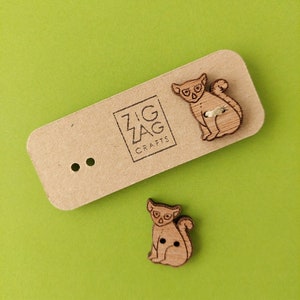 Könnte beinhalten: Zwei Holz-Knöpfe mit einem Cartoon-Lemuren-Design. Die Knöpfe sind braun und haben schwarze Augen und eine schwarze Nase. Die Knöpfe befinden sich auf einer braunen Karte mit dem Text "Zig Zag Crafts".