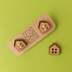 Puede incluir: Tres botones de madera en forma de casa con un acabado de madera natural. Dos de los botones tienen un pequeño trozo de cuerda tejido a través de los agujeros. Los botones están en una tarjeta marrón con el texto "Zig Zag Crafts".