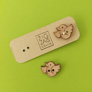 May include: Two wooden buttons shaped like birds with brown wings and a light brown body. The buttons have two holes each and are on a brown card with the text "Zig Zag Crafts".