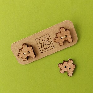 May include: Three wooden puzzle piece buttons with a natural wood finish. The buttons are on a brown card with the text "Zig Zag Crafts".