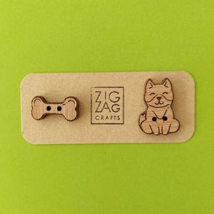 May include: Two wooden buttons, one shaped like a bone and the other with a cartoon dog design. The buttons are on a brown card with the text "Zig Zag Crafts".