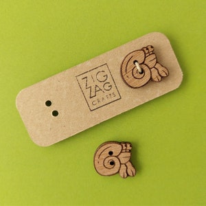 May include: Two wooden buttons shaped like hermit crabs. The buttons are brown and have a natural wood finish. The buttons are on a brown card with the text "Zig Zag Crafts" printed on it.