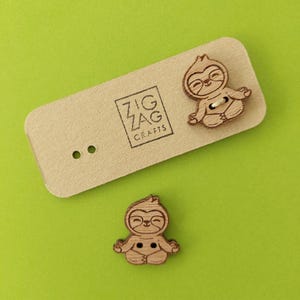 May include: Two wooden buttons shaped like sloths in a yoga pose. The buttons are brown and have a white string attached to them. The buttons are on a brown card with the text "Zig Zag Crafts" printed on it.