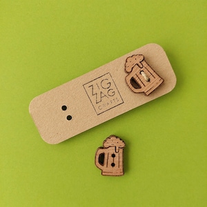 May include: Two wooden beer mug buttons and a rectangular tag with the text "ZIG ZAG CRAFTS" printed on it. The buttons are light brown with etched details, and the tag is a light brown color. The background is a solid green.