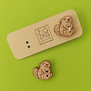 Puede incluir: Dos botones de madera con forma de castor, con una cara sonriente y sosteniendo un palo. Los botones están en una tarjeta marrón con el texto "Zig Zag Crafts".