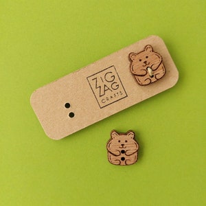 May include: Two wooden hamster-shaped buttons with a light brown color and etched details. One button is attached to a brown tag with the text "ZIG ZAG CRAFTS". The background is a solid green.
