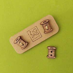 Puede incluir: Tres botones de madera en forma de carrete de hilo, con un acabado de madera marrón. Los botones tienen forma de carrete de hilo y tienen un agujero en el centro. Los botones están en una tarjeta marrón con el texto "Zig Zag Crafts" impreso en ella.