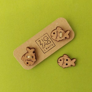 May include: Three wooden fish buttons with a simple design. The buttons are on a brown card with the text "Zig Zag Crafts".