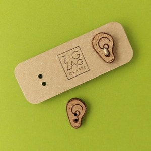 Può includere: Due orecchini di legno a forma di orecchie. Gli orecchini sono su una carta marrone con il testo "Zig Zag Crafts" stampato su di essa.