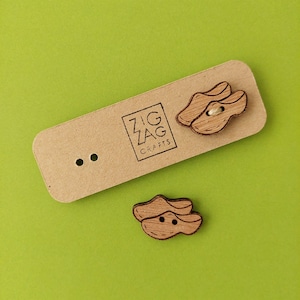 May include: Two wooden buttons shaped like clogs, one pair is on a brown card with the text "Zig Zag Crafts", the other pair is on a green surface.