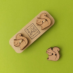 Op de afbeelding: Drie houten knopen in de vorm van eekhoorns. De knopen zijn bruin en hebben een natuurlijke houten afwerking. De knopen bevinden zich op een bruine kaart met de tekst "Zig Zag Crafts" erop gedrukt.