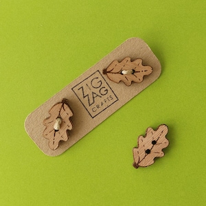 May include: Three wooden oak leaf buttons with a brown stain. The buttons are on a brown card with the text "Zig Zag Crafts".