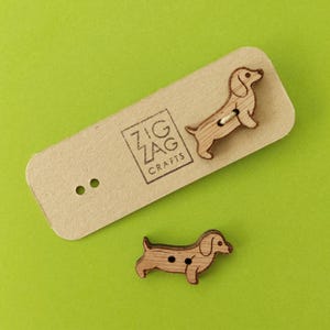 Könnte beinhalten: Zwei Holz-Knöpfe in Form von Dackeln. Die Knöpfe sind braun und haben eine natürliche Holzoptik. Der größere Knopf befindet sich auf einer Karte mit dem Text "Zig Zag Crafts".