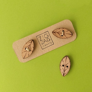 May include: Three wooden leaf-shaped buttons with a natural wood finish. The buttons have a small hole in the centre and are displayed on a brown card with the text "Zig Zag Crafts".