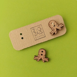 Può includere: Due bottoni di legno a forma di polpo di colore legno marrone. I bottoni sono su una carta marrone con il testo "Zig Zag Crafts".