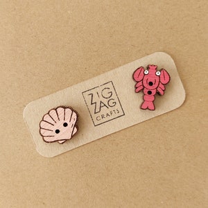 May include: A set of two wooden buttons featuring a pink seashell and a pink lobster with googly eyes. The buttons are displayed on a brown card with the text "Zig Zag Crafts".