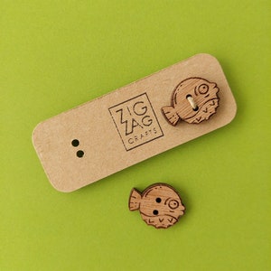 Peut inclure: Deux boutons en bois en forme de poisson-globe avec une finition en bois brun. Les boutons sont sur une carte brune avec le texte "Zig Zag Crafts".