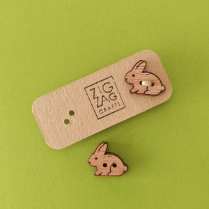 Puede incluir: Dos botones de madera con forma de conejo con una mancha marrón. Los botones están sobre una tarjeta marrón con el texto "Zig Zag Crafts" impreso en ella.