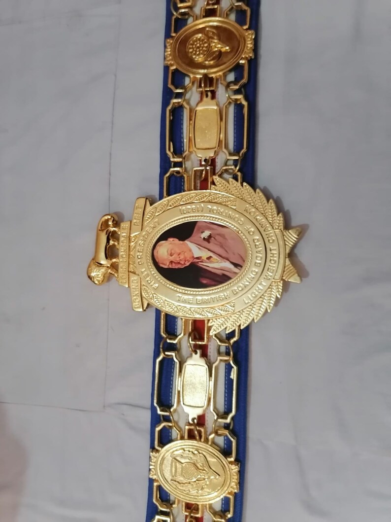 May include: A gold boxing championship belt with a blue and red strap. The belt features a large oval medallion with a portrait of a man and the text "THE BRITISH BOXING BOARD OF CONTROL LIGHTWEIGHT CHAMPION 1969/1974".