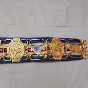 May include: A gold belt with blue and white ribbon and three gold medallions. The medallions feature a flower, a scene with two people, and a flower.