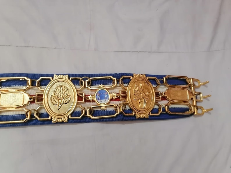 May include: A gold belt with blue and white ribbon and three gold medallions. The medallions feature a flower, a scene with two people, and a flower.