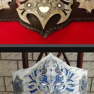 Könnte beinhalten: Zwei Meisterschaftsgürtel mit einer zentralen Metallplatte in Form einer stilisierten Maske. Der obere Gürtel hat einen braunen Lederriemen mit goldfarbenen Metallplatten, der untere Gürtel einen weißen Riemen mit silber- und blaufarbenen Metallplatten.