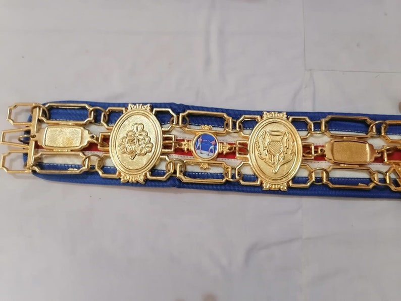 May include: A gold belt with a blue and white ribbon. The belt has three oval medallions with different designs. The medallions feature a clover, a thistle, and a scene of two people fencing.