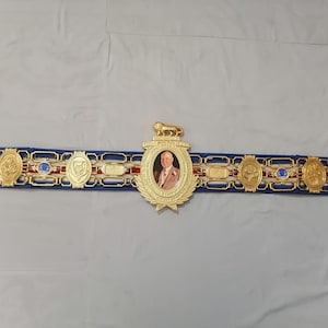 May include: A gold boxing championship belt with a blue strap and red and white accents. The belt features a large central medallion with a portrait of a man in a suit and tie. The medallion is surrounded by smaller medallions with various designs.