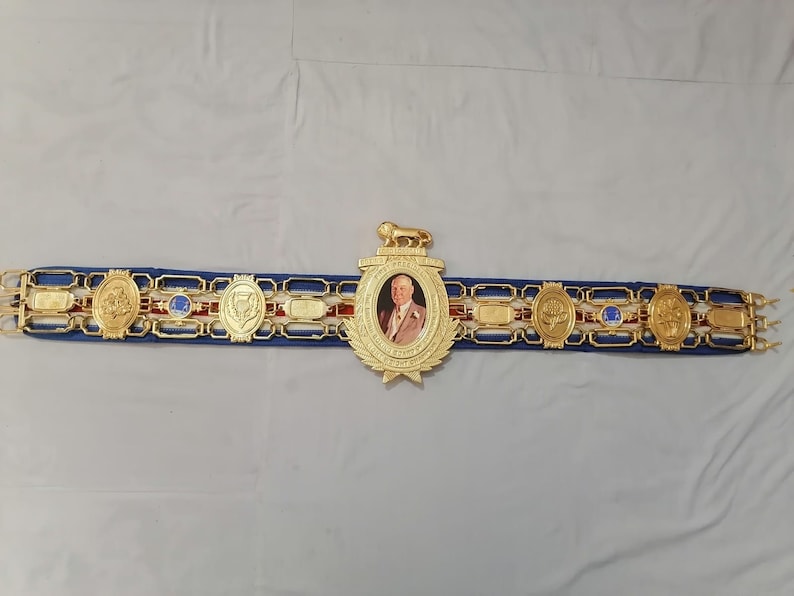 May include: A gold boxing championship belt with a blue strap and red and white accents. The belt features a large central medallion with a portrait of a man in a suit and tie. The medallion is surrounded by smaller medallions with various designs.