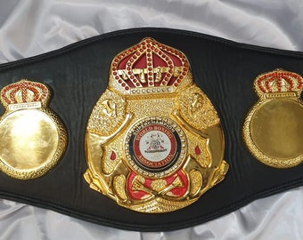 May include: A black leather boxing championship belt with gold and red accents. The centre plate features a crown, two lions, and a circular emblem with the text "World Boxing Association". The belt has two side plates with crowns and blank spaces for engraving.