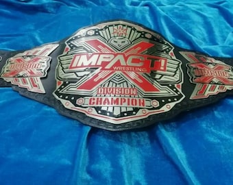 Può includere: Una cintura da campionato di wrestling in pelle nera con accenti argentati e rossi. La cintura presenta le parole "Impact! Wrestling" e "Campione di divisione".