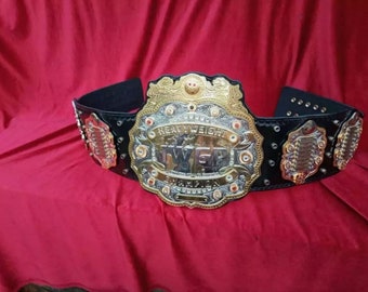 Op de afbeelding: Een gouden en zilveren kampioensgordel met zwarte leren banden en metalen accenten. De gordel heeft de woorden "Heavyweight" en "IWGP" in gouden letters.