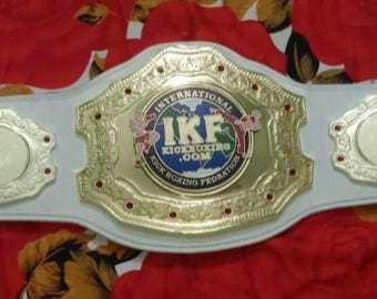 May include: A white championship belt with gold accents. The belt features a large, circular emblem with the text "International Kickboxing Federation" and "IKF" in the center. The belt is on a red and white floral background.