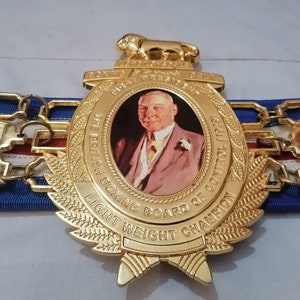 May include: A gold boxing championship belt with a lion head emblem and a portrait of a man. The belt reads "The British Boxing Board of Control 1921" and "Light Weight Champion".