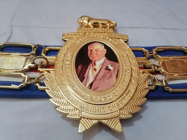 May include: A gold boxing championship belt with a lion head emblem and a portrait of a man. The belt reads "The British Boxing Board of Control 1921" and "Light Weight Champion".