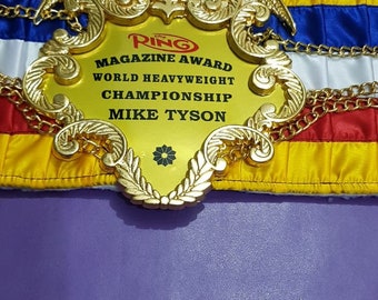 Op de afbeelding: Een gouden bokskampioensgordel met een adelaarontwerp en de tekst "The Ring Magazine Award World Heavyweight Championship Mike Tyson".