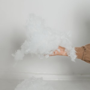 May include: A hand holds a cloud of fluffy, white polyester fiberfill. The fiberfill is soft and voluminous, with a light and airy appearance. The background is a plain white wall and floor.