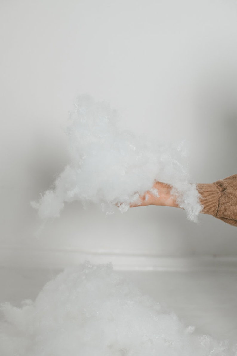 May include: A hand holds a cloud of fluffy, white polyester fiberfill. The fiberfill is soft and voluminous, with a light and airy appearance. The background is a plain white wall and floor.