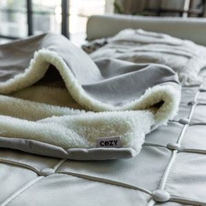 May include: A gray blanket with a white, fluffy interior is draped over a white leather couch. The blanket has a tag that reads "COZY". The blanket's soft texture and neutral color palette suggest comfort and relaxation.