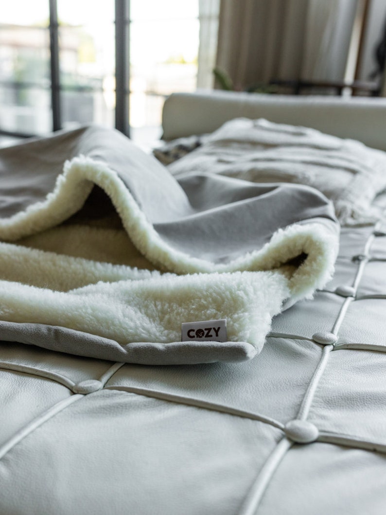 May include: A gray blanket with a white, fluffy interior is draped over a white leather couch. The blanket has a tag that reads "COZY". The blanket's soft texture and neutral color palette suggest comfort and relaxation.