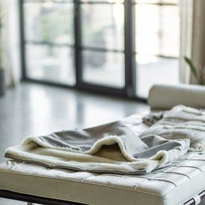 May include: A gray blanket with a cream-colored, fluffy lining is draped over a white leather bench with a dark brown frame. The blanket is folded, revealing the soft interior. The scene is set in a modern, well-lit room.