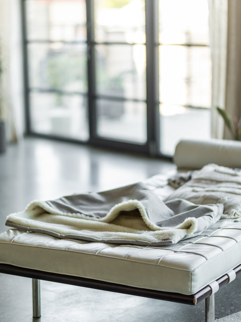 May include: A gray blanket with a cream-colored, fluffy lining is draped over a white leather bench with a dark brown frame. The blanket is folded, revealing the soft interior. The scene is set in a modern, well-lit room.