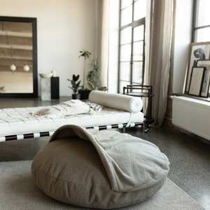 Pu&ograve; includere: Un letto rotondo per animali domestici, dai toni neutri, con un design a cappuccio, &egrave; posto su un tappeto grigio chiaro. Il letto &egrave; il punto focale in una stanza luminosa e ariosa, con un divano letto in pelle bianca e grandi finestre.