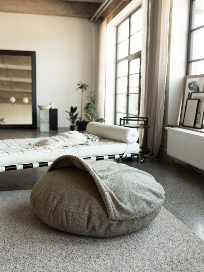 Pu&ograve; includere: Un letto rotondo per animali domestici, dai toni neutri, con un design a cappuccio, &egrave; posto su un tappeto grigio chiaro. Il letto &egrave; il punto focale in una stanza luminosa e ariosa, con un divano letto in pelle bianca e grandi finestre.